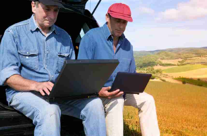 Two farmers researching land properties online with laptops in a field.