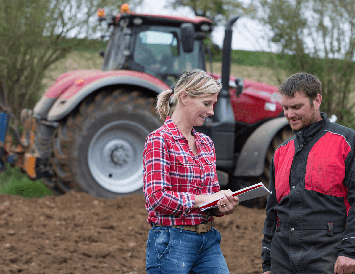Male and female farmer discussing while there's a red tractor in background.