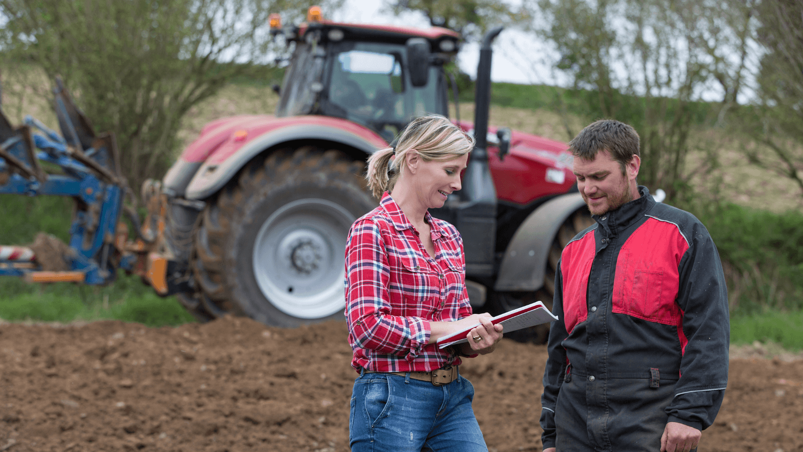 Farming Couple Talking About List Alt Text: Male and female farmer discussing while there's a red tractor in background.