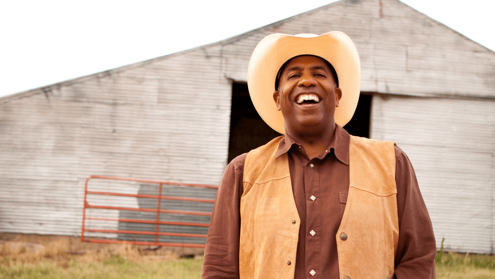 Black Cowboy With Huge Smile On Face Alt Text: Contented black male farmer with huge barn in the background.