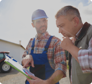 Two farmers in casual work attire discuss plans while one holds a clipboard with documents, standing outdoors near farming equipment under bright sunlight.