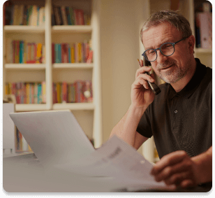 A man in glasses works from home, speaking on the phone while reviewing documents in front of a laptop, with bookshelves in the background.