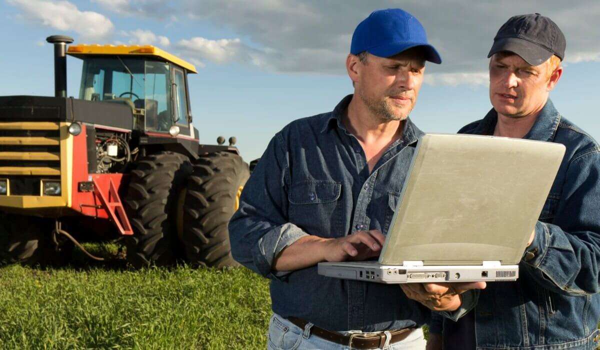 Two male farmers holding a laptop in an open field. There's a tractor in the background.