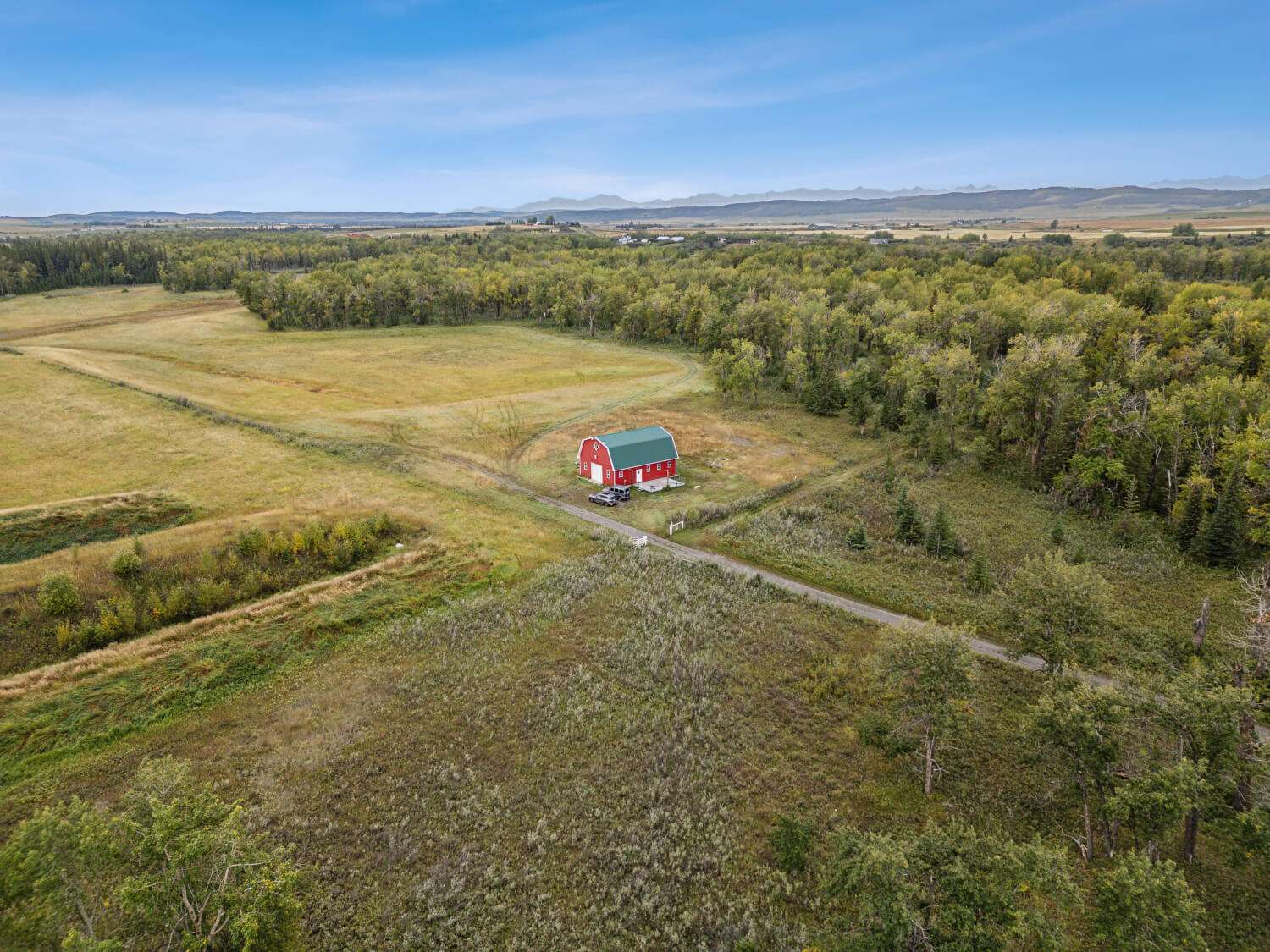 A red barn sits alone in an open countryside field, surrounded by trees, with distant mountains under a bright blue sky.