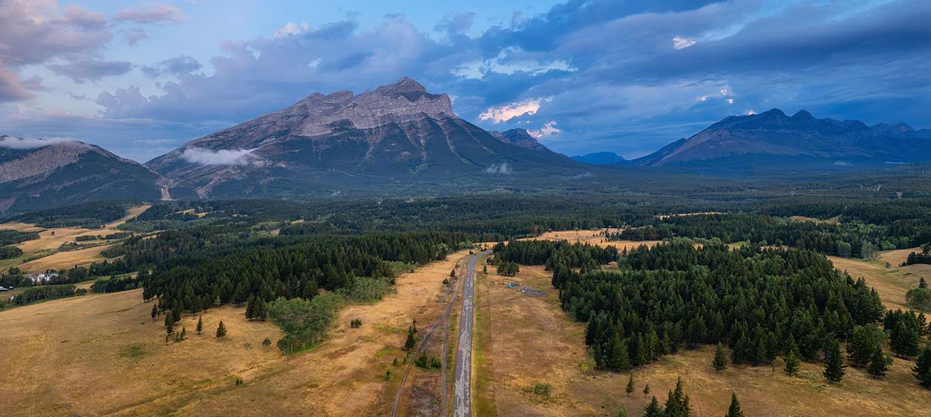 A stunning aerial view of a winding road cutting through open fields and dense forest, with towering mountains in the background under a soft, cloudy dusk sky.