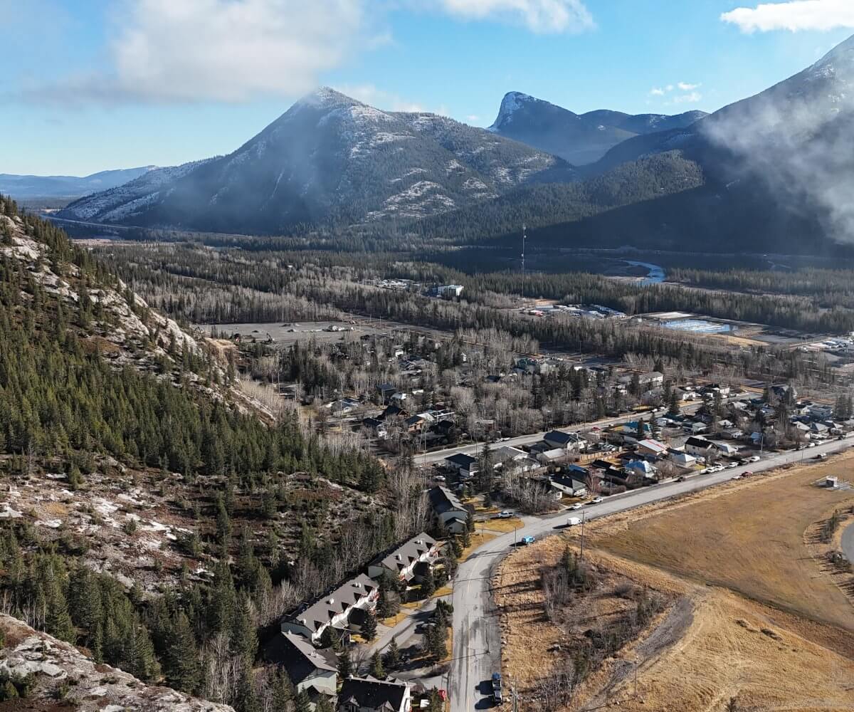 Aerial view of a small mountain town in Alberta with forested slopes, a winding river valley, and snow-dusted peaks under low cloud