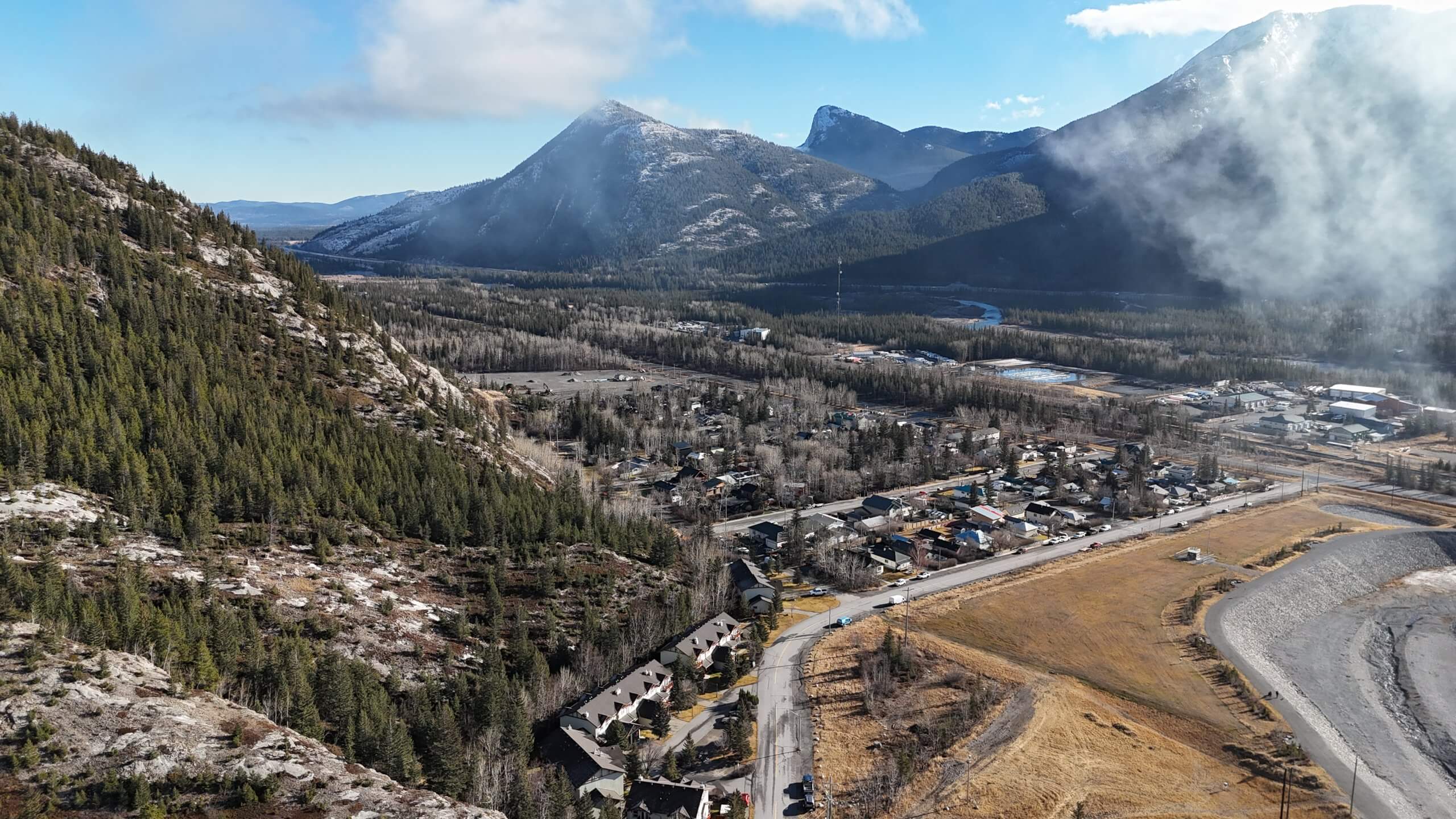 Aerial view of a small mountain town in Alberta with forested slopes, a winding river valley, and snow-dusted peaks under low cloud